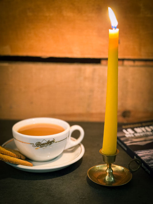 A yellow beeswax taper andle and tea cup on a table with a warm glow