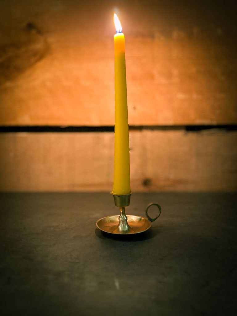 A yellow beeswax taper candle in a brass holder against a textured wall.