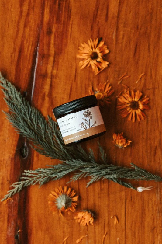 A brown glass jar with a flowered label "Crème à mains" on top of a wood table with a fir branch and some calendula flowers