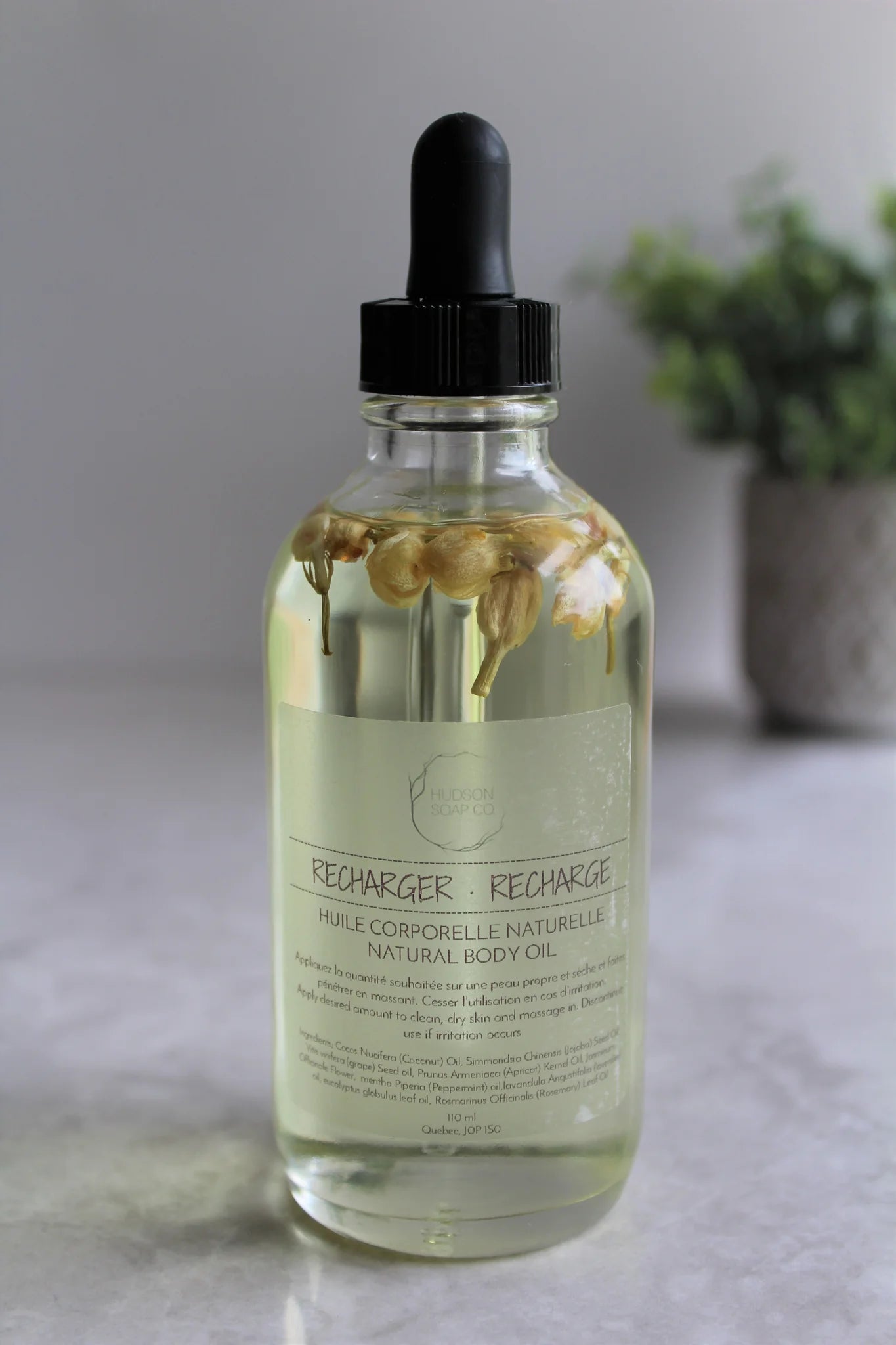 Bottle of Clear natural body oil with flower buds suspended in oil "Recharge"  with a plant off focus in background