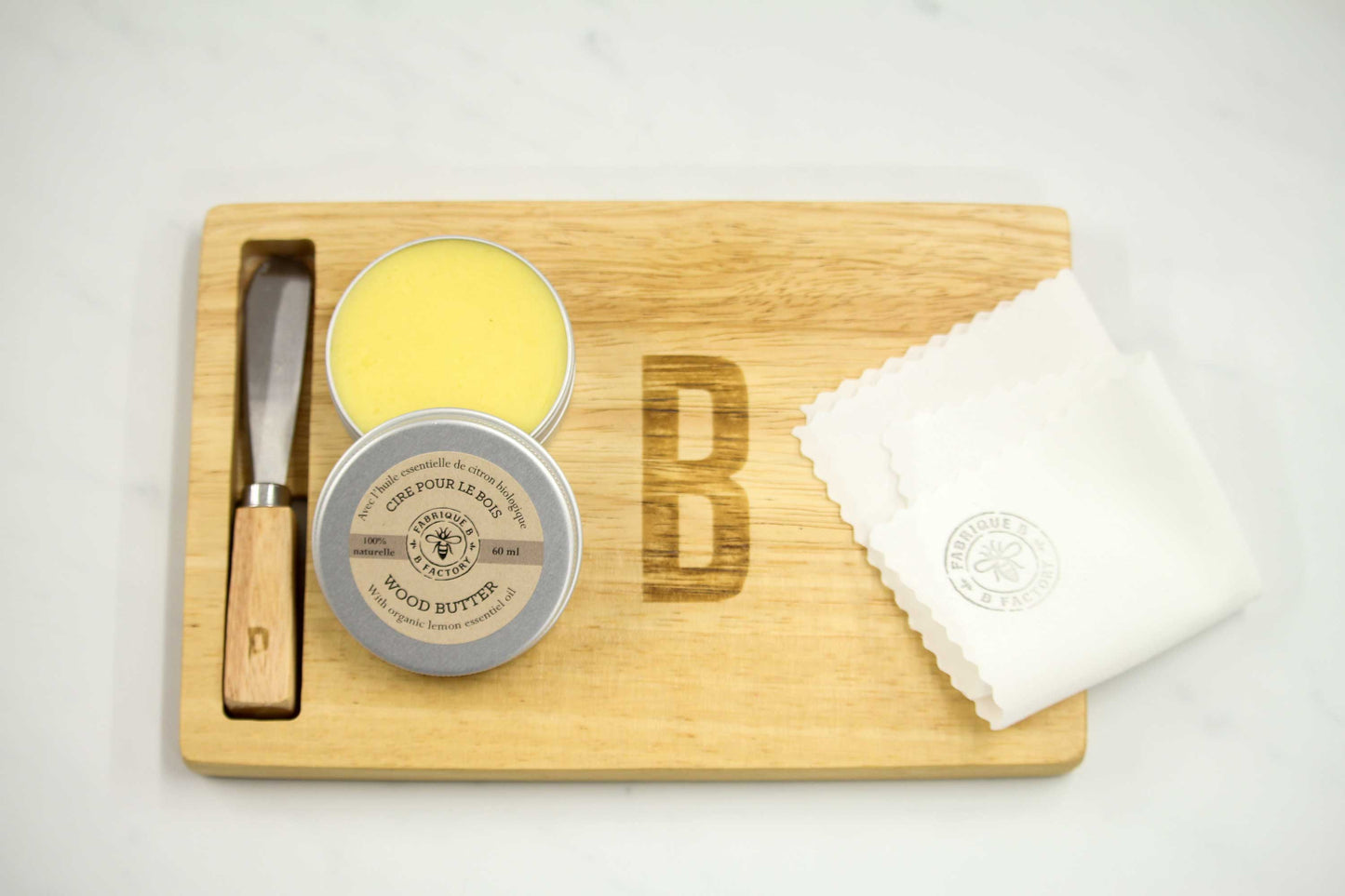 B Factory natural beeswax wood butter in open silver tin next to white cloth on small wood charcuterie board