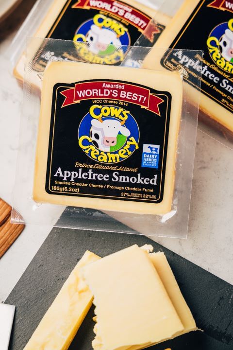 A block of Cows Creamery raw milk apple-smoked cheddar cheese, featuring a firm texture with a rich color and subtle smoky aroma in branded packaging.
