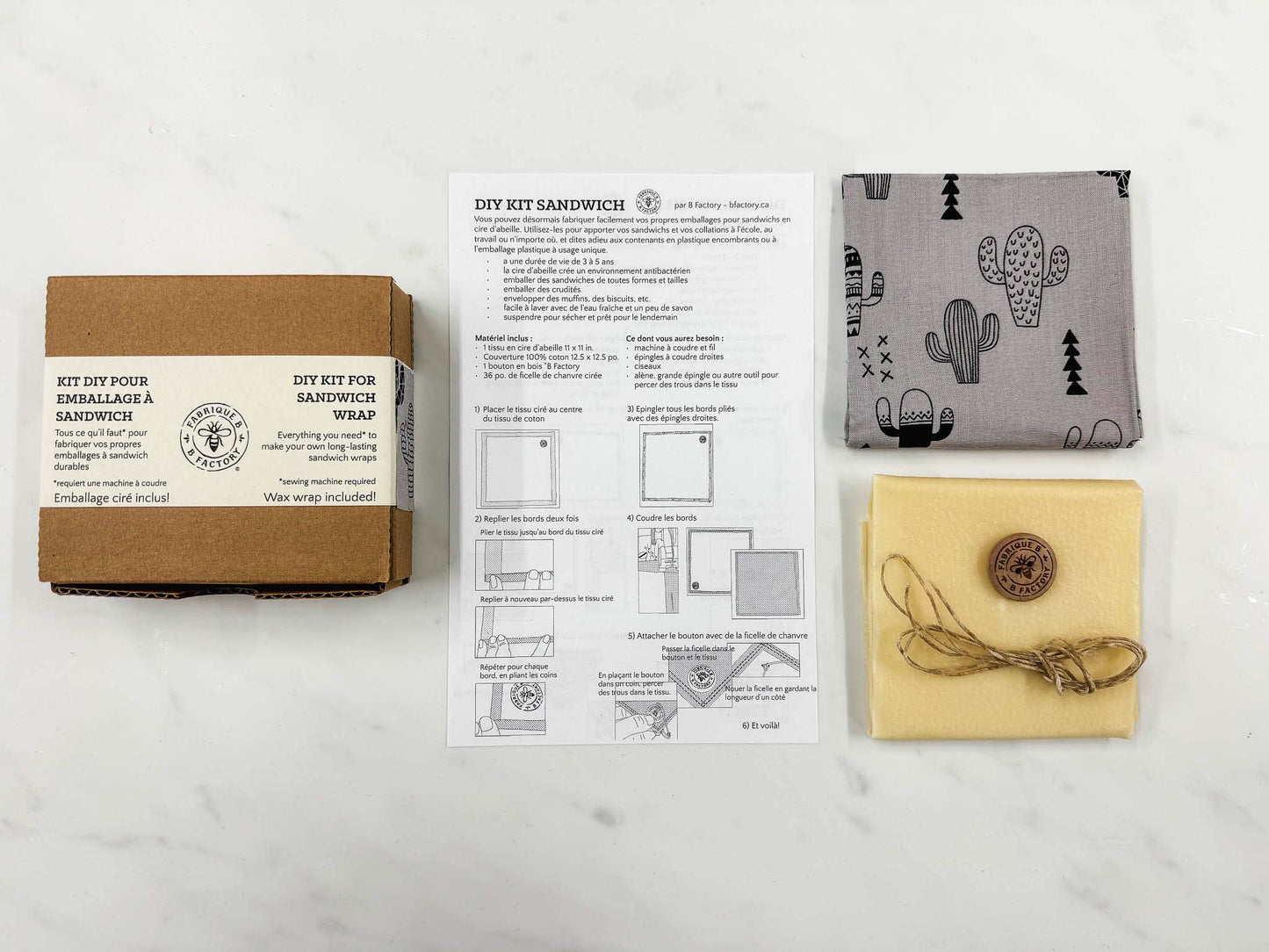 A cardboard box of a KIY kit for sandwich wrap, a set of instructions, a piece of grey fabric with a cactus pattern, a beeswax wrap, a button and a waxed hemp twine all set on a white background