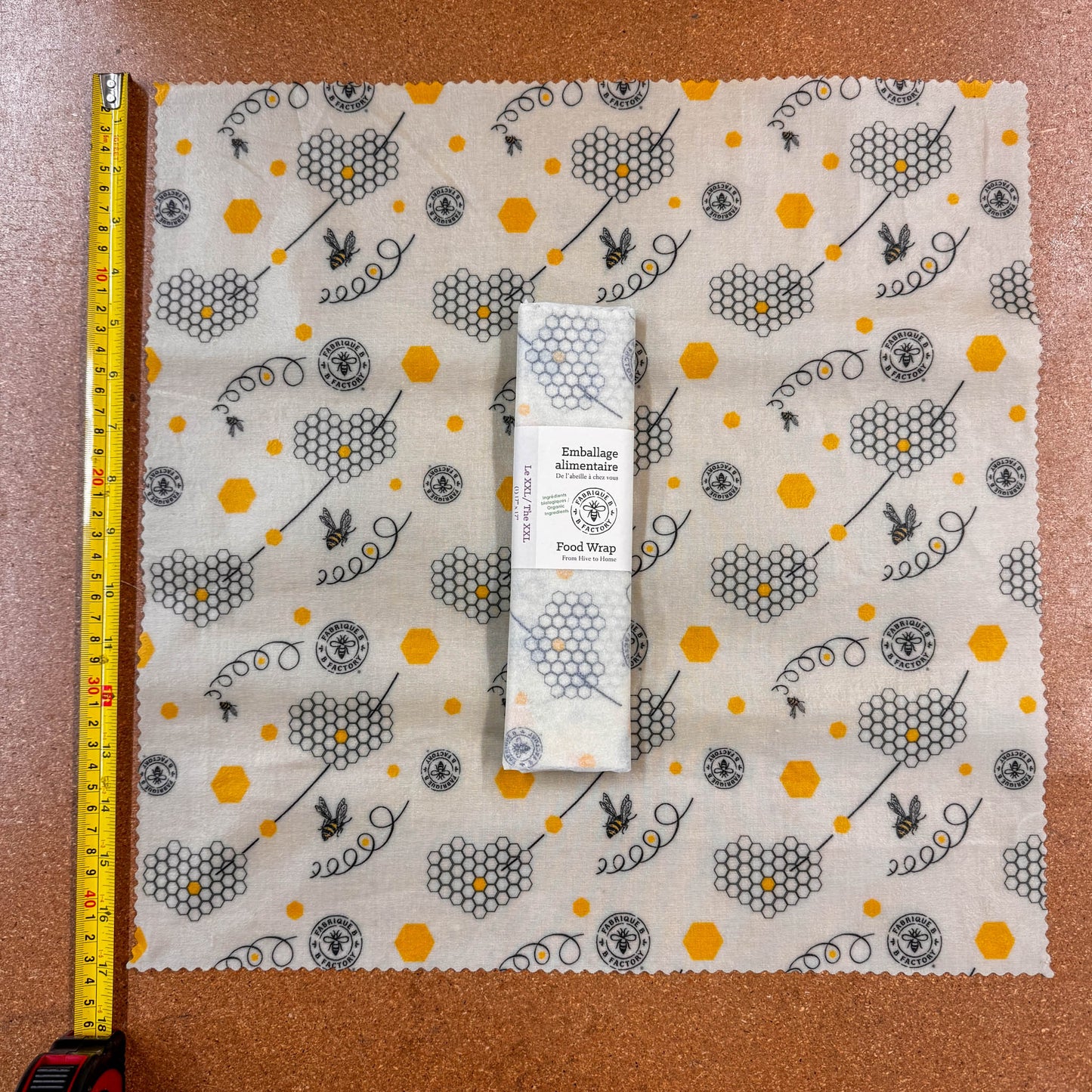 B Factory beeswax foodwrap with honeycomb and bee pattern, folded and placed on a brown surface with a measuring tape along the side for size comparison.