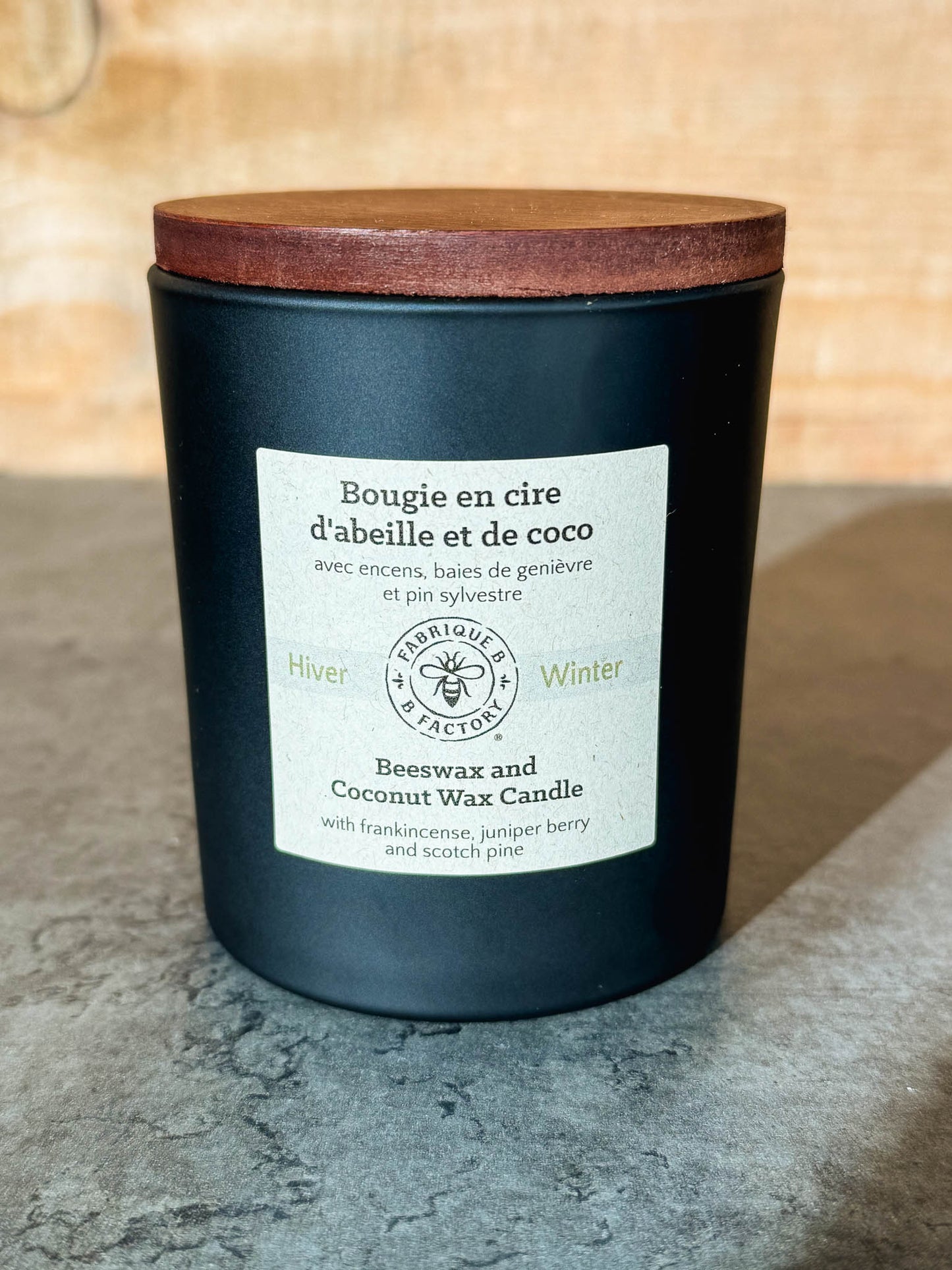 B Factory winter beeswax and coconut wax candle with scotch pine and juniper berry in a black jar and wood lid, sitting on a grey counter with a wood background