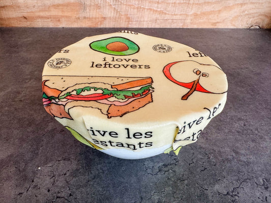 B Factory small beeswax food wrap covering a bowl with "i love leftovers/vive les restants" from Laucolo printed on the wrap with half an apple,  half-eaten sandwich, and half an avocado also in the design