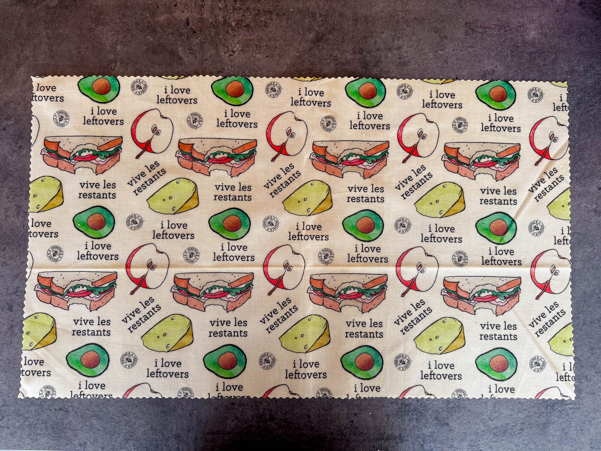 B Factory giant beeswax foodwrap with "i love leftovers/vive les restants" from Laucolo printed on the wrap with half an apple,  half-eaten sandwich, and half an avocado also in the design