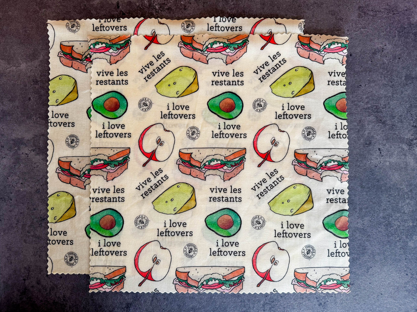 B Factory two large beeswax food wraps on a grey counter with "i love leftovers/vive les restants" from Laucolo printed on the wrap with half an apple,  half-eaten sandwich, and half an avocado also in the design