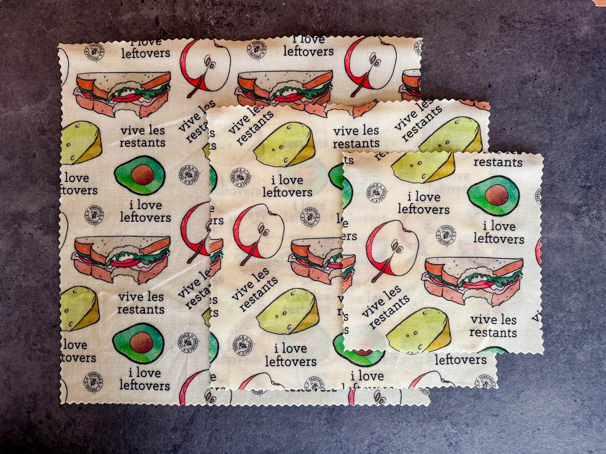 B Factory beeswax food wraps - one small, one medium, and one large - on a grey counter with "i love leftovers/vive les restants" from Laucolo printed on the wrap with half an apple,  half-eaten sandwich, and half an avocado also in the design