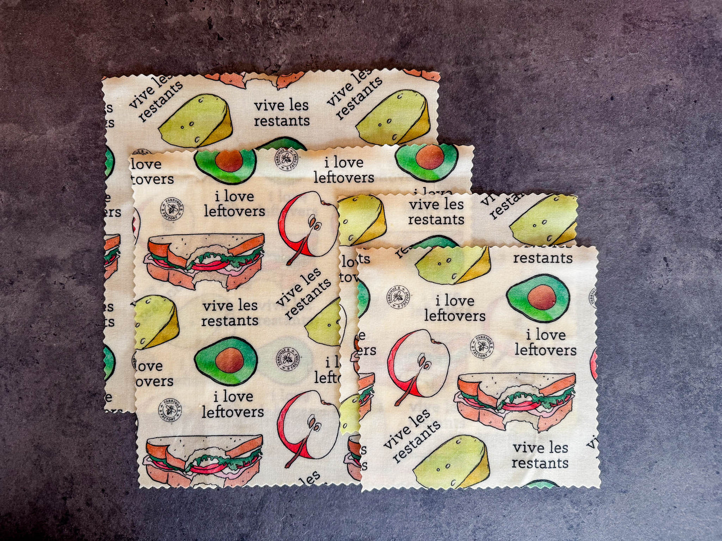 B Factory beeswax food wraps - two small, two medium - on a grey counter with "i love leftovers/vive les restants" from Laucolo printed on the wrap with half an apple,  half-eaten sandwich, and half an avocado also in the design