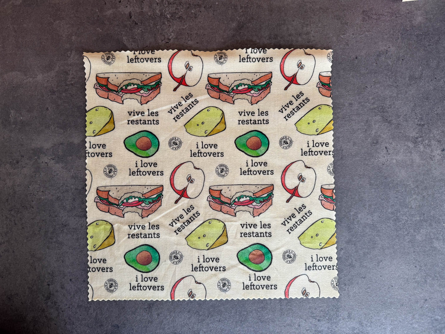 B Factory beeswax food wrap - one large - on a grey counter with "i love leftovers/vive les restants" from Laucolo printed on the wrap with half an apple,  half-eaten sandwich, and half an avocado also in the design