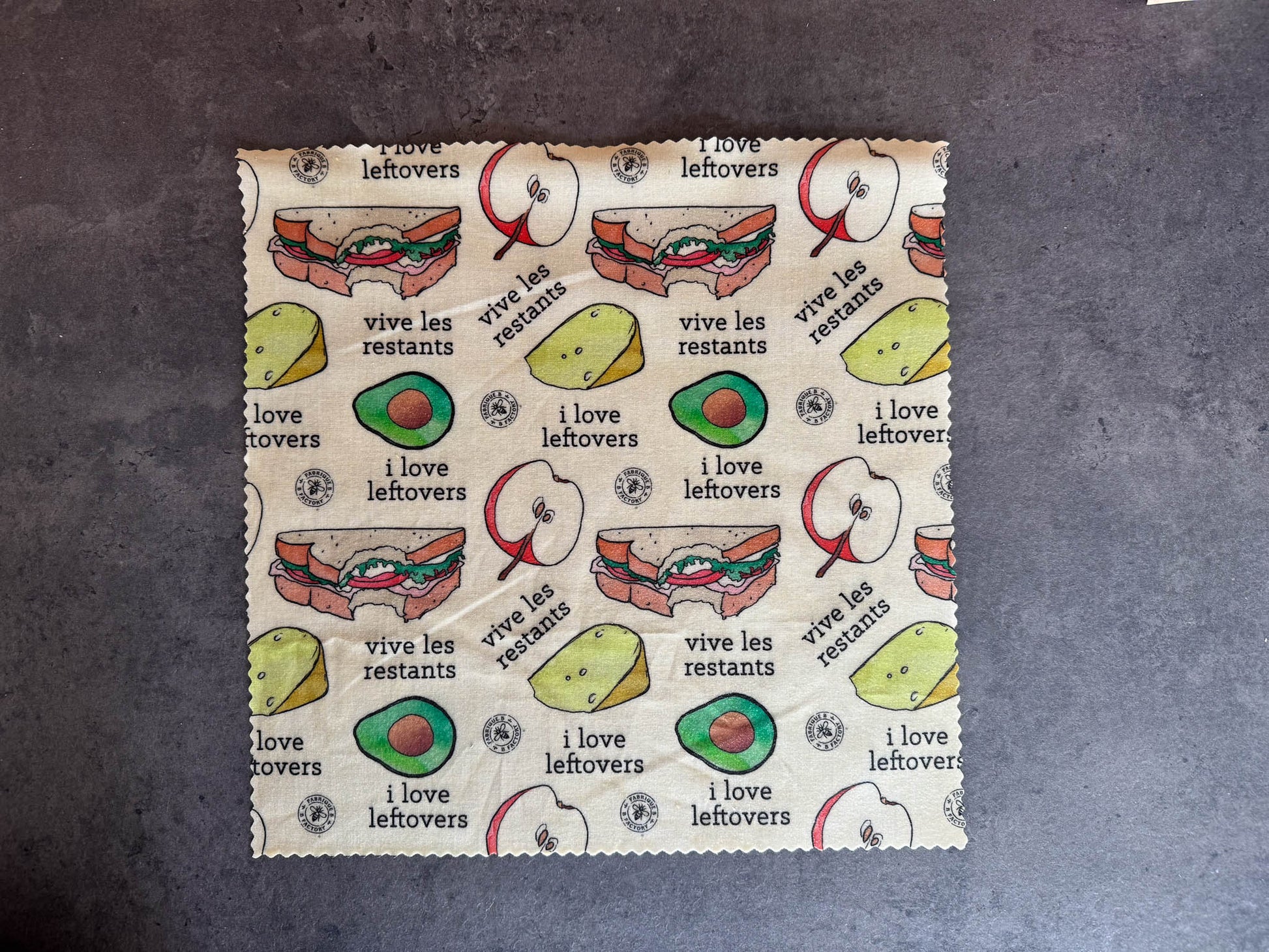 B Factory beeswax food wrap - one large - on a grey counter with "i love leftovers/vive les restants" from Laucolo printed on the wrap with half an apple,  half-eaten sandwich, and half an avocado also in the design