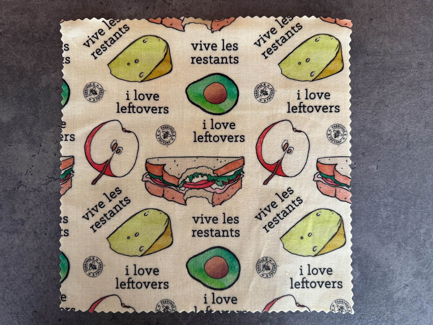 B Factory beeswax food wraps - one medium - on a grey counter with "i love leftovers/vive les restants" from Laucolo printed on the wrap with half an apple,  half-eaten sandwich, and half an avocado also in the design