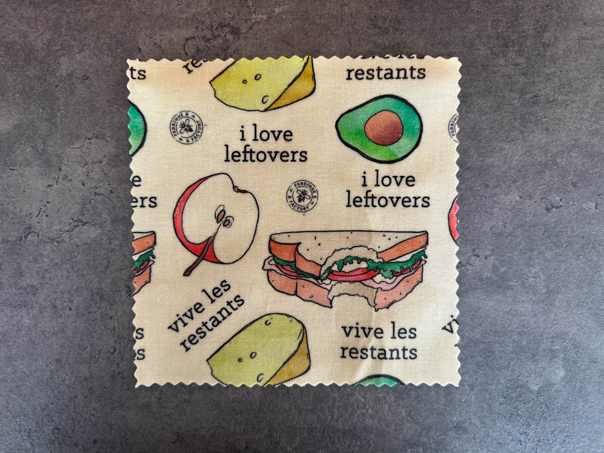 B Factory beeswax food wraps - one small - on a grey counter with "i love leftovers/vive les restants" from Laucolo printed on the wrap with half an apple,  half-eaten sandwich, and half an avocado also in the design