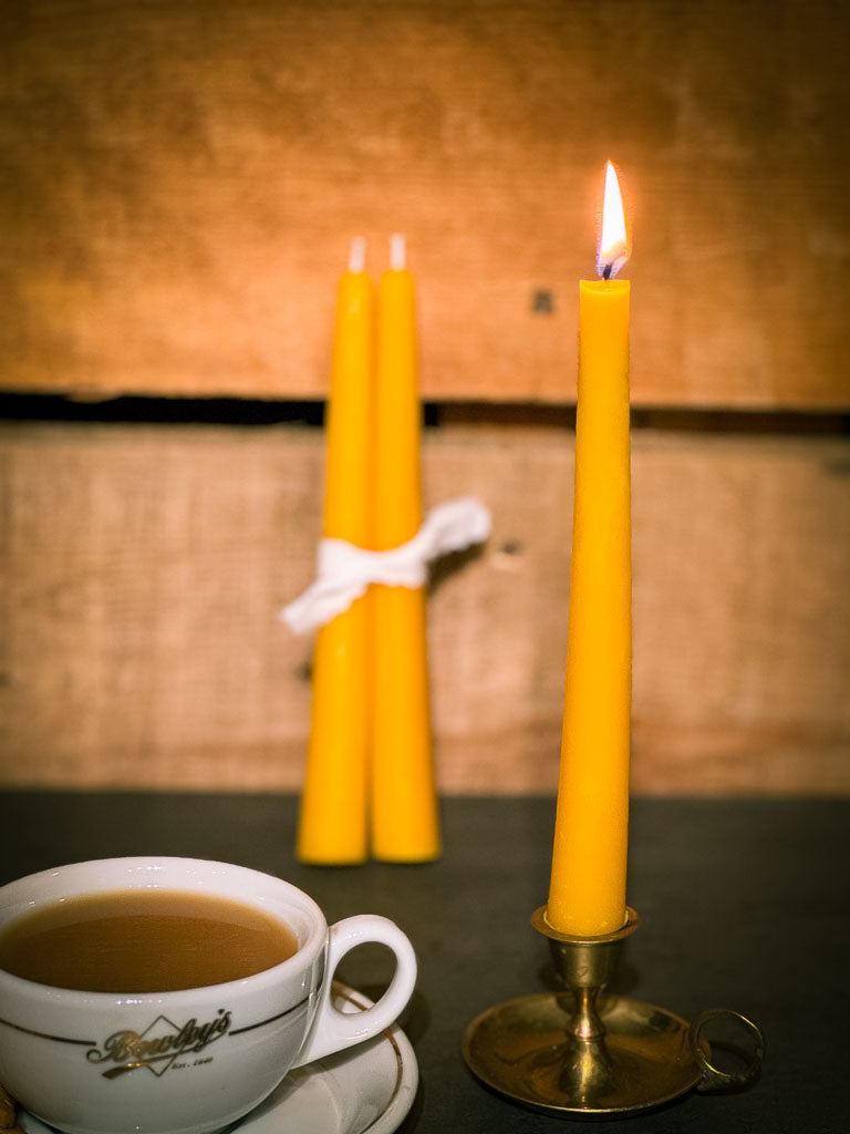 A yellow beeswax taper candle in a brass candlestick with a cup of coffee on a wooden surface, and a pair of beeswax taper candles in background.