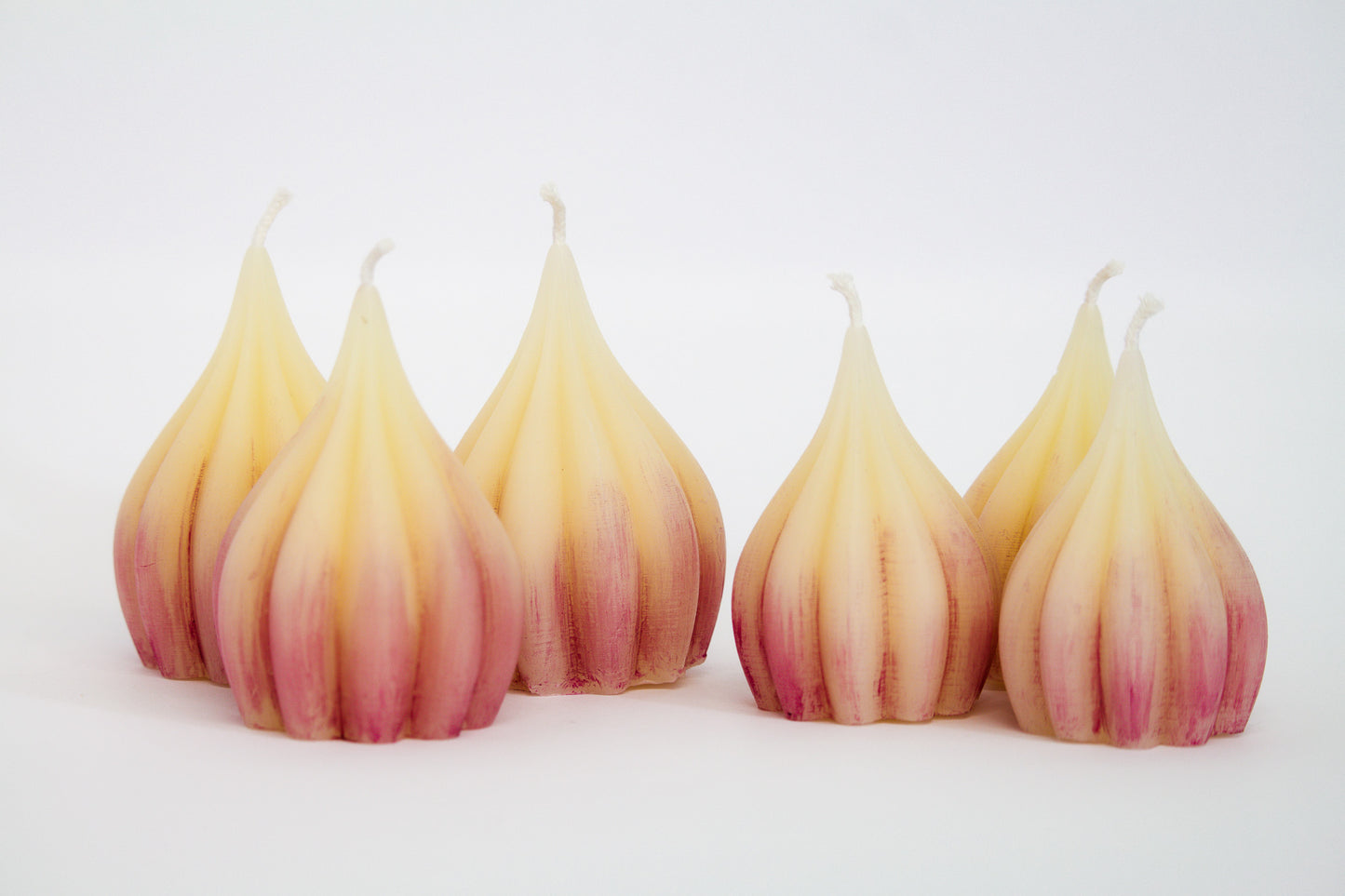 Garlic-shaped white beeswax candles painted with some purple. Three large on left and three smaller ones on right
