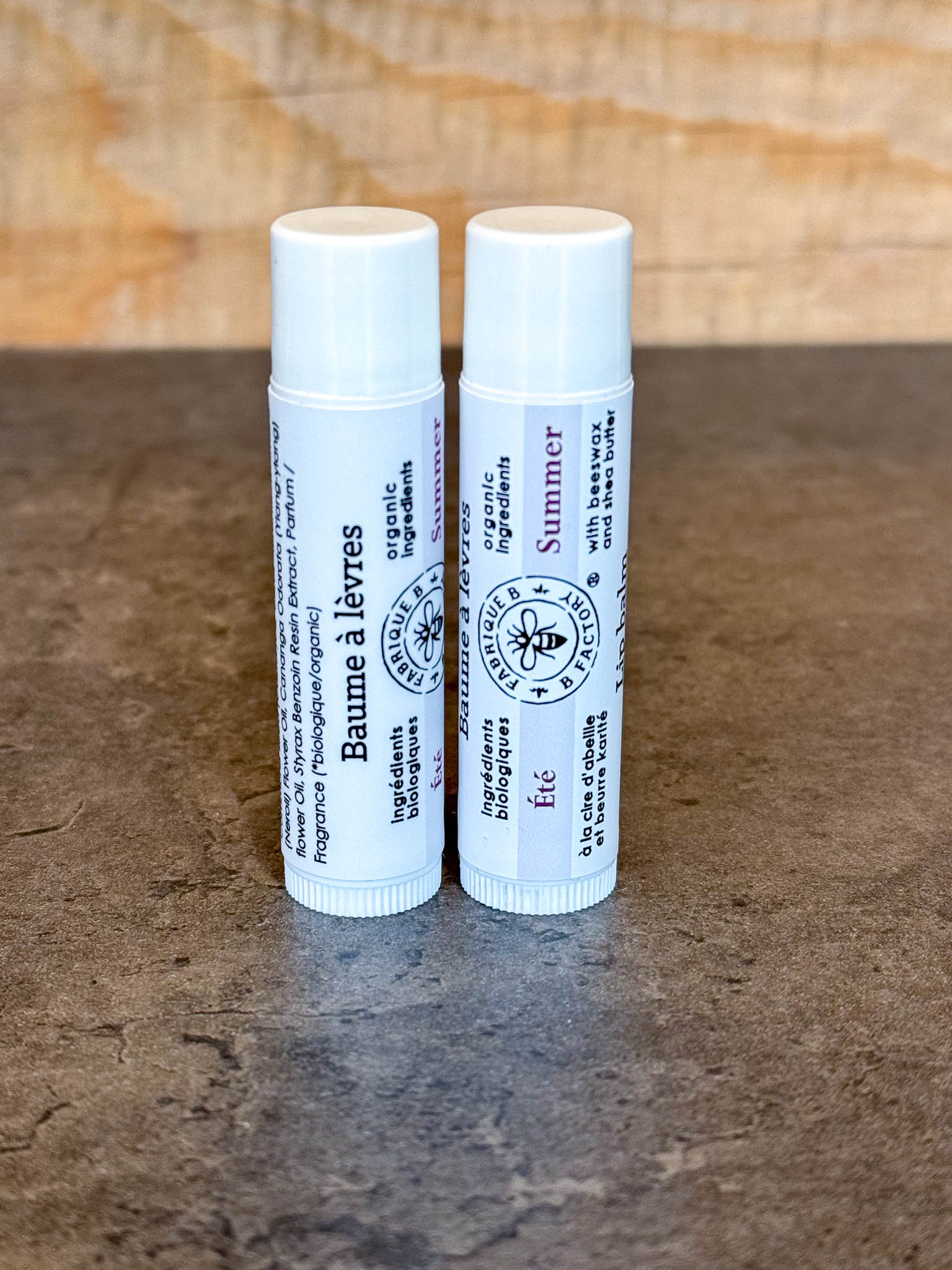 2 "Summer" lip balms by B Factory standing on end, on a dark surface with a wood background