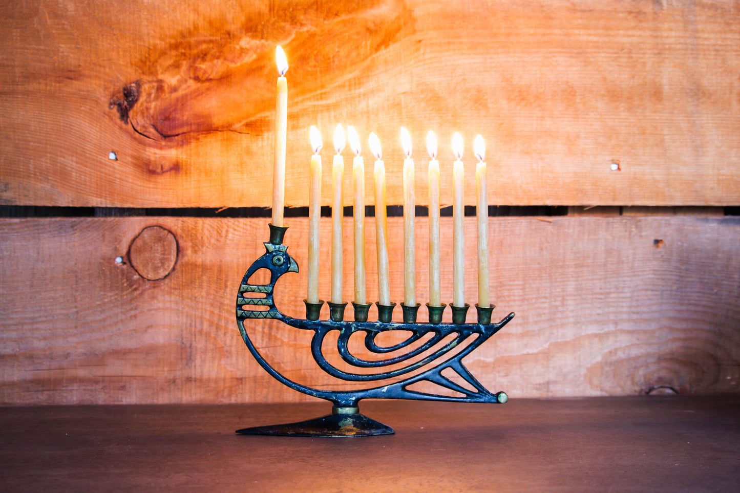 9 lit pure beeswax candles on an antique menorah shaped like a bird, on a grey countertop with a wood background