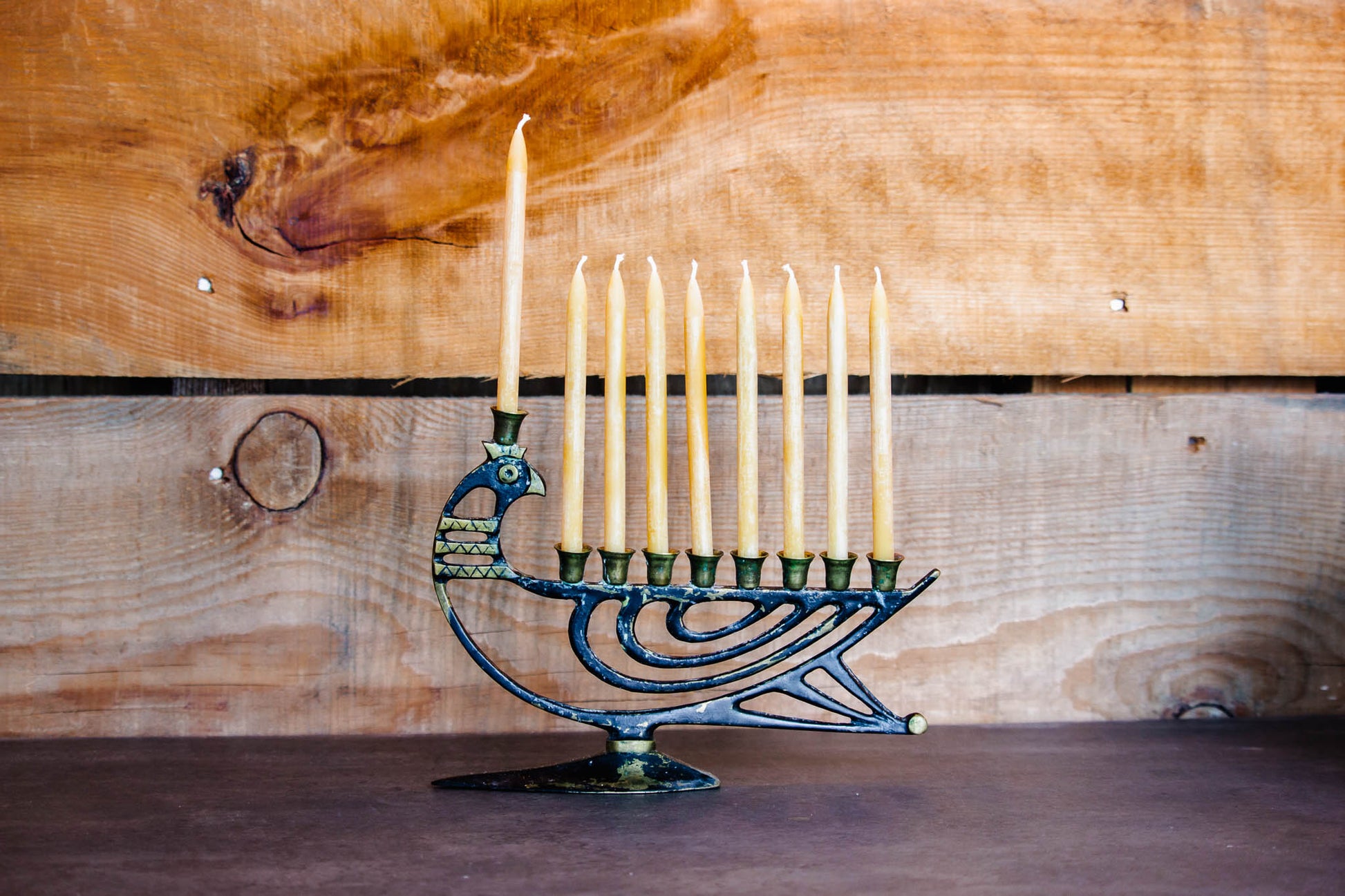 9 unlit pur beeswax candles on an antique menorah shaped like a bird, on a grey countertop with a wood background