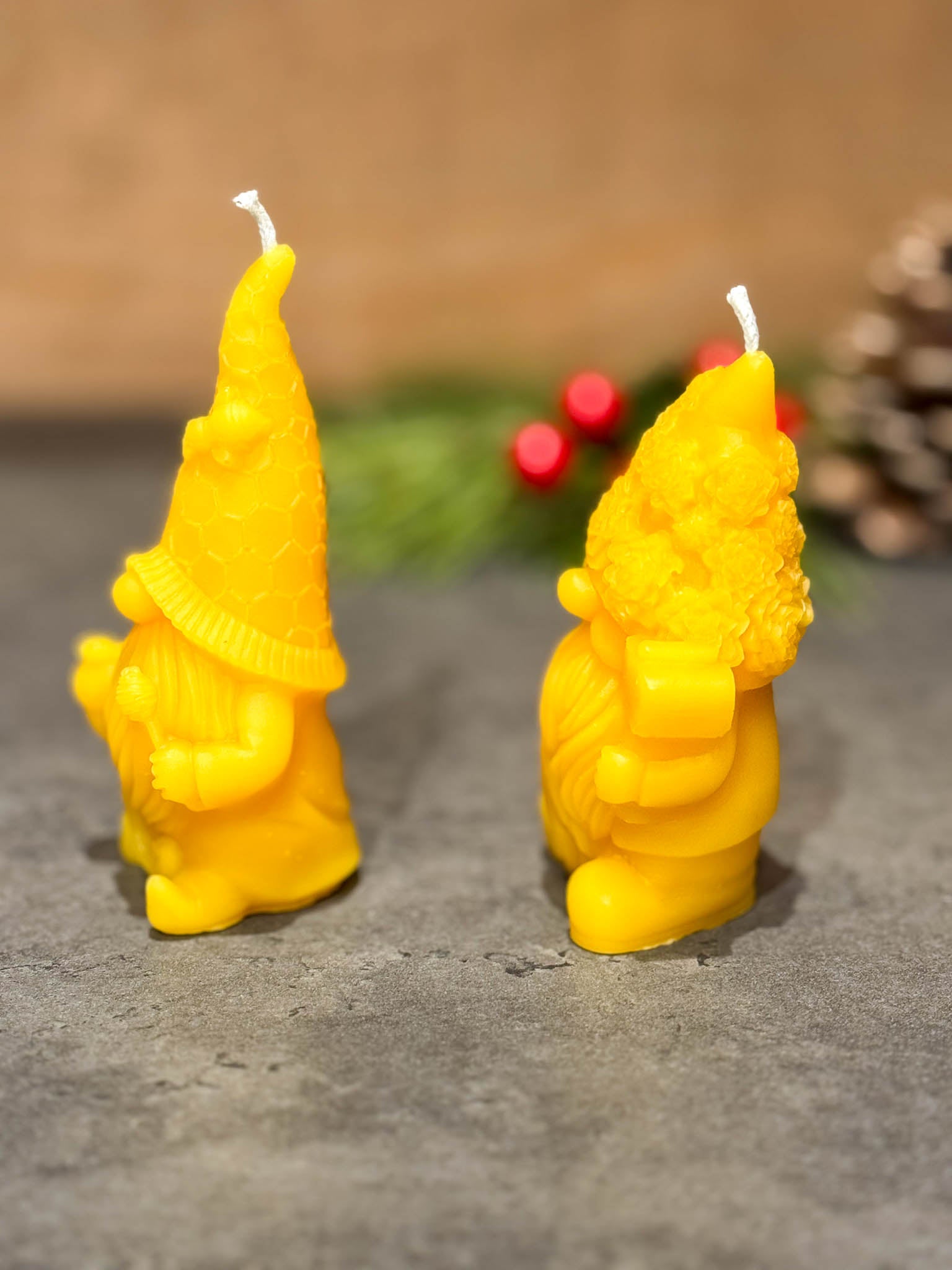 Two pure beeswax gnome candles on a grey counter with green branches with berries in the background - seen from the left side