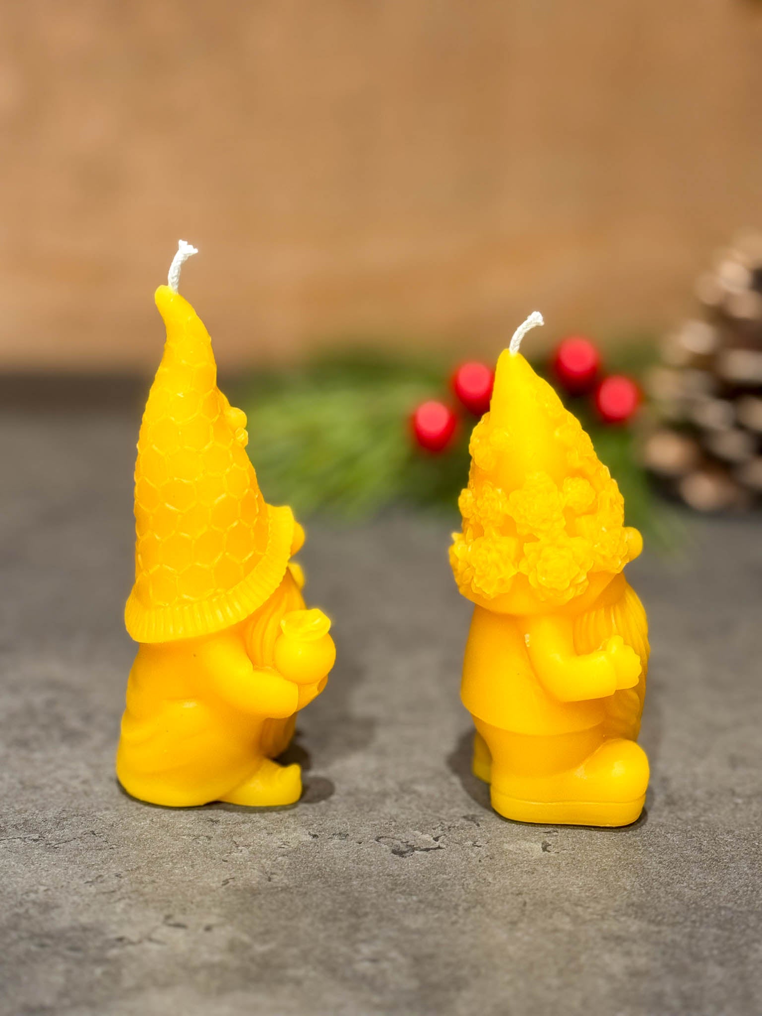 Two pure beeswax gnome candles on a grey counter with green branches with berries in the background - seen from the right side