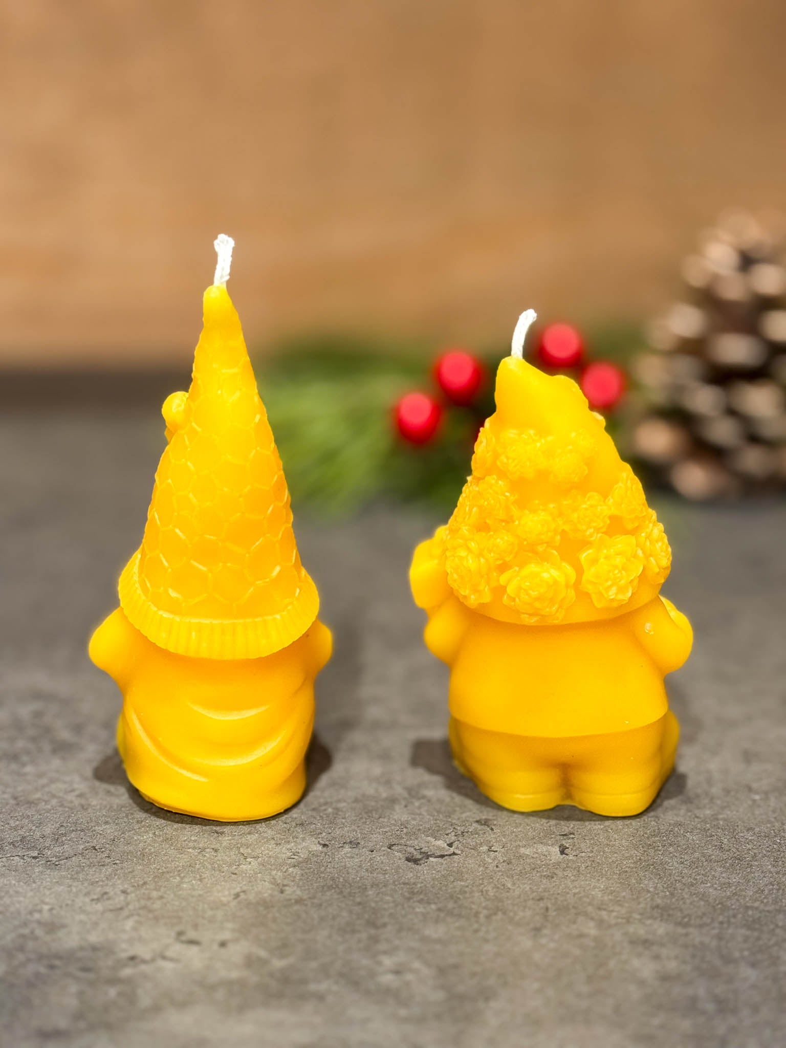 Two pure beeswax gnome candles on a grey counter with green branches with berries in the background - seen from the back