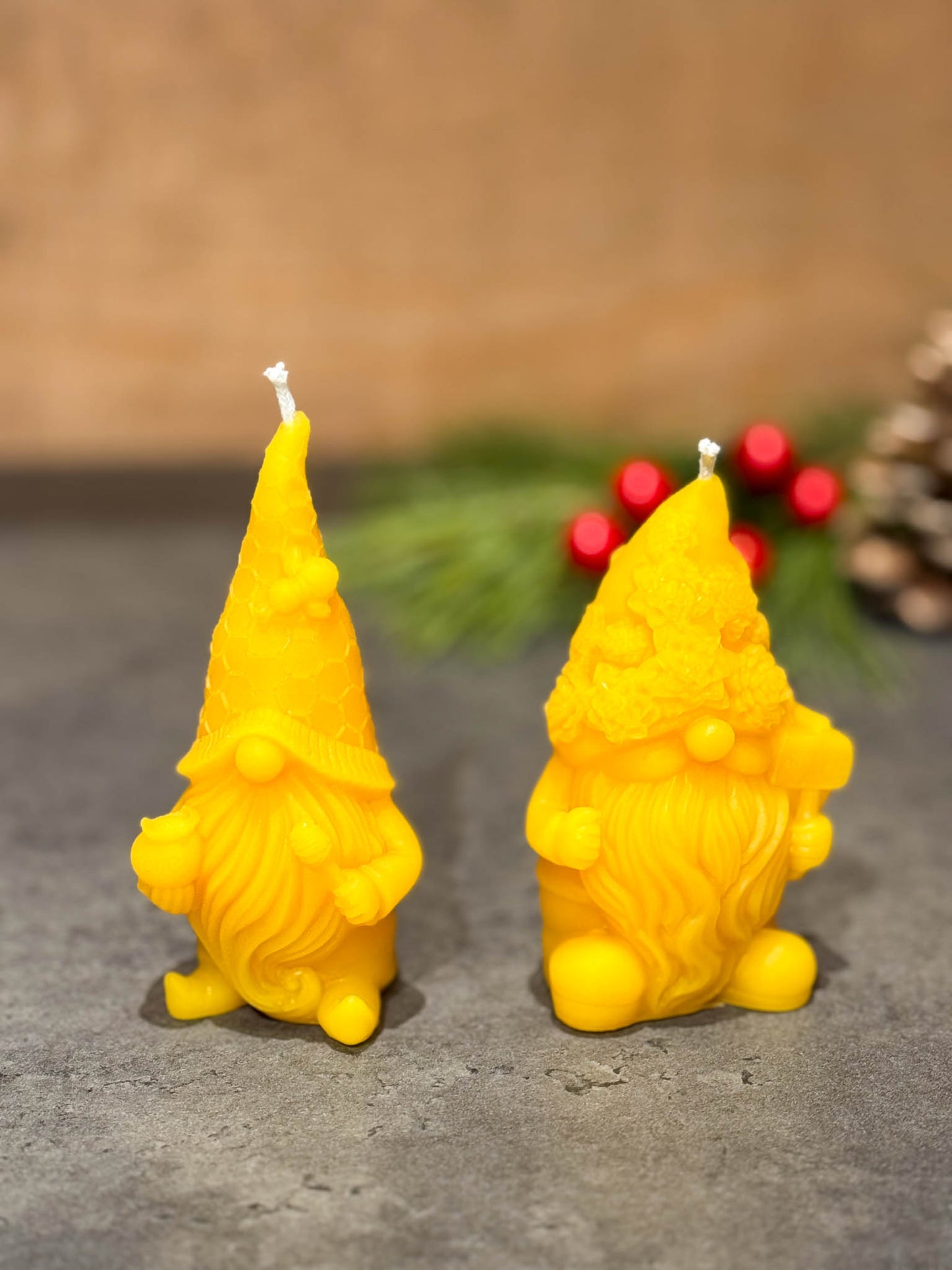 Two pure beeswax gnome candles on a grey counter with green branches with berries in the background - seen from the front