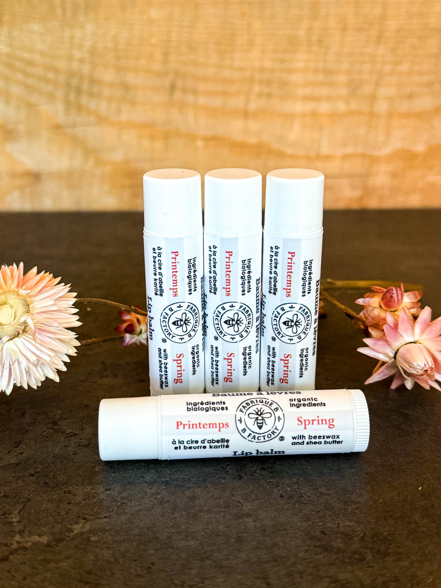 3 "Spring" lip balms by B Factory standing on end, with another lip balm lying in front, on a dark surface with a wood background and some dried flowers on the side