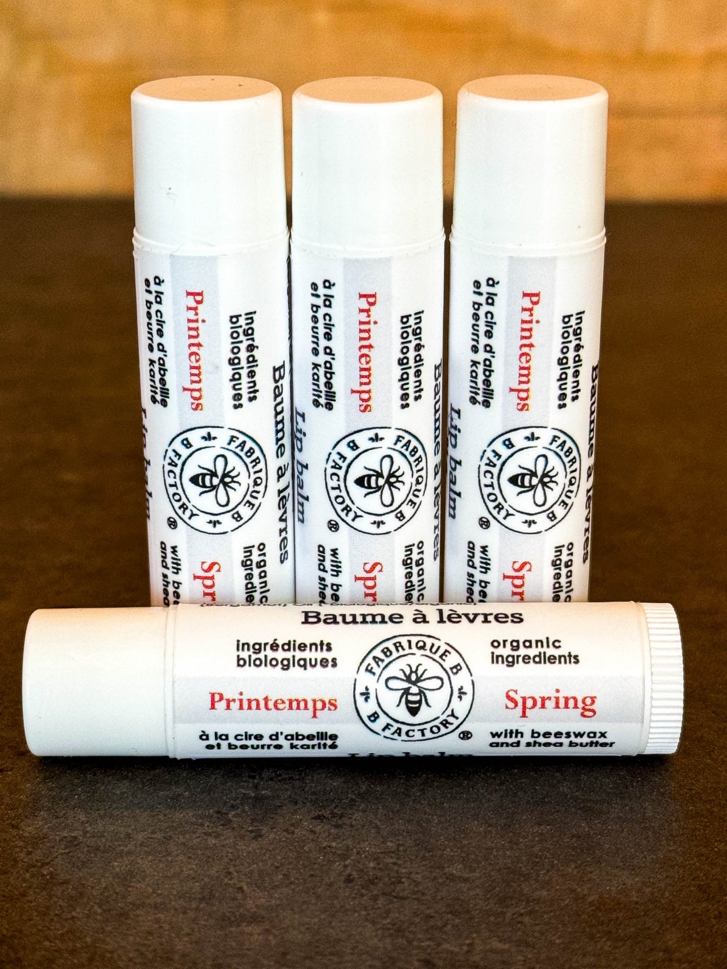 3 "Spring" lip balms by B Factory standing on end, with another lip balm lying in front, on a dark surface with a wood background