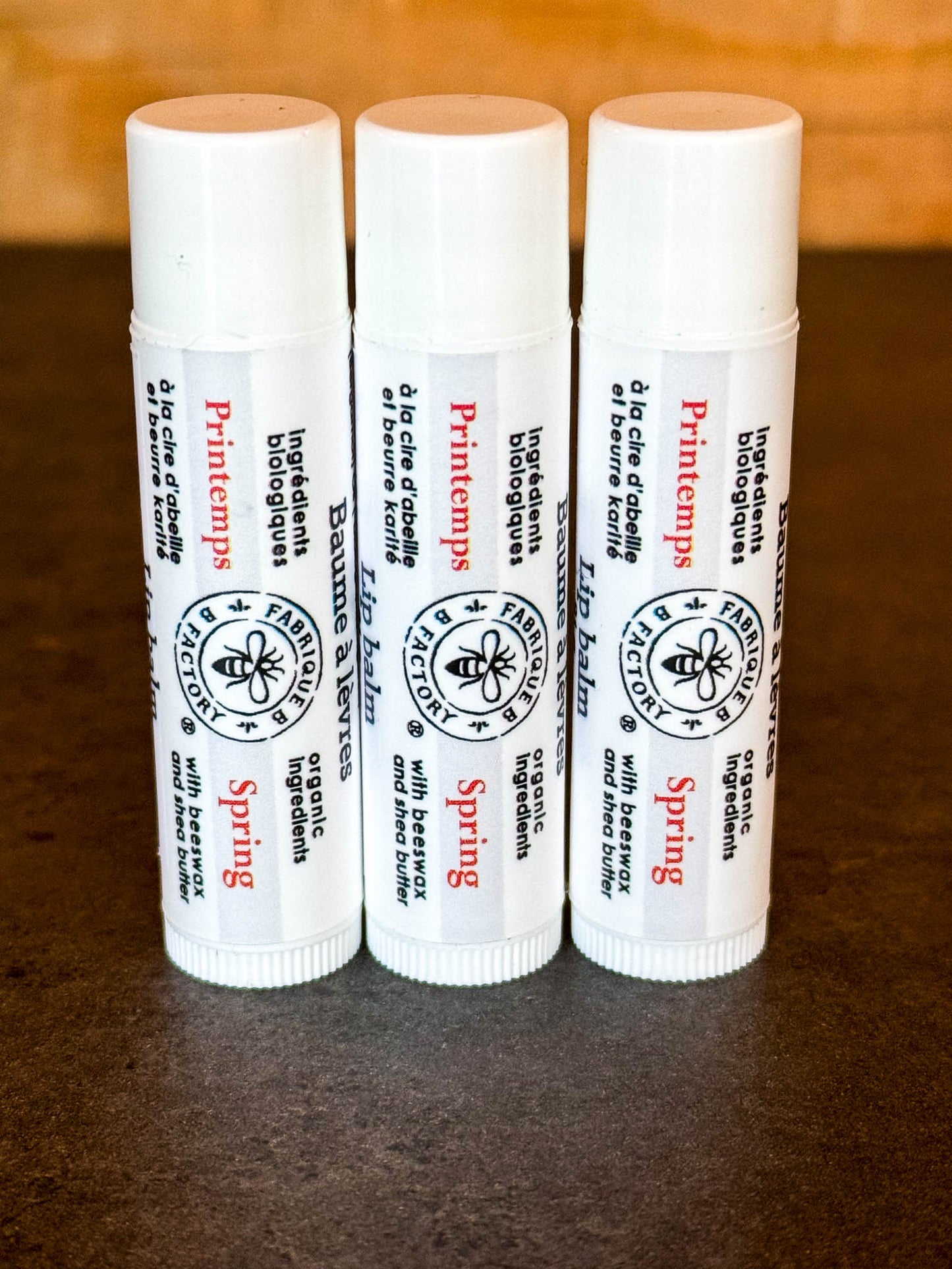 3 "Spring" lip balms by B Factory standing on end, on a dark surface with a wood background