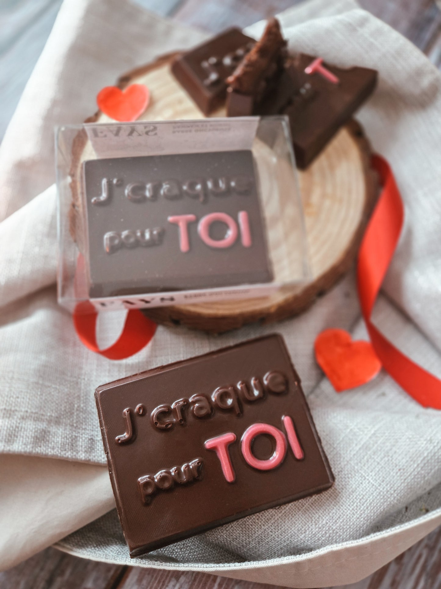 A milk chocolate bar engraved with “J’craque pour TOI” displayed with heart decorations, ribbon, and a gift box, styled for a romantic occasion.