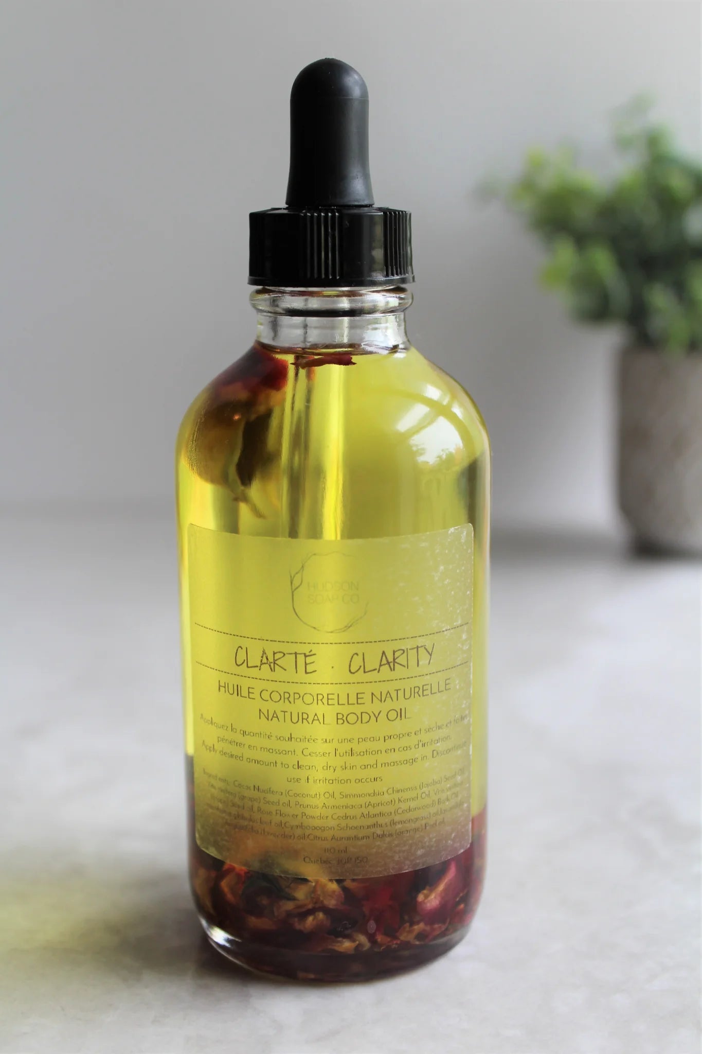 Bottle of natural body oil with red flowers suspended in the oil "Clarity" with a plant off focus in background