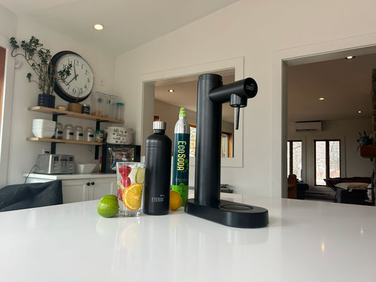 Black soda dispenser with EcoSoda Co2 canister and Eterna black stainless steel bottle on a kitchen counter with drinks and fruit