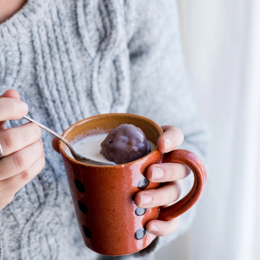 Person holding a brown mug with a spoon, containing a dark chocolate melting bomb in hot milk, against a neutral background.