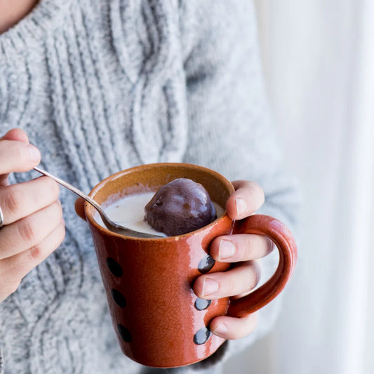Person holding a brown mug with a spoon, containing a dark chocolate melting bomb in hot milk, against a neutral background.