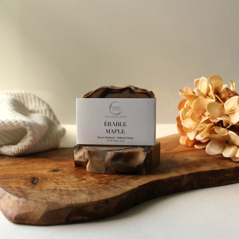 "Maple" bar soap by Hudson Soap Co. set on a wood plank, with orange flowers to the right and a soft white cloth to the left, on a white background