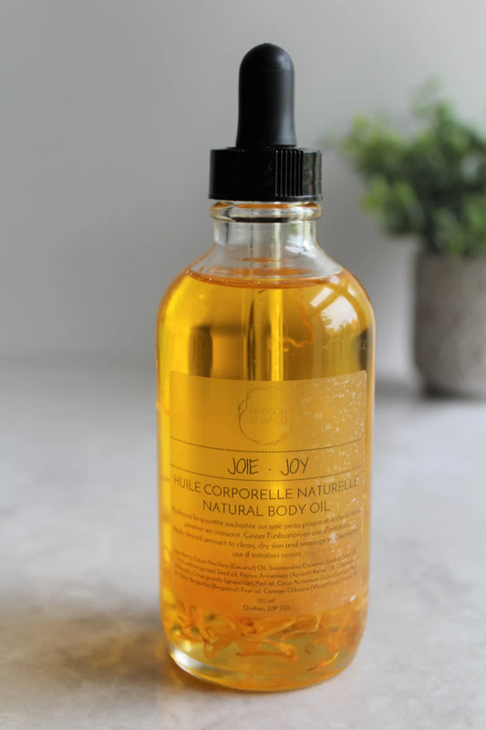 Bottle of golden coloured body oil "Joy" with a plant off focus in background