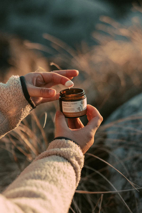 One hand is holding an open jar of "crème à mains", while the other hand has one finger with some of the cream on the tip. Background blurred of dry grasses.