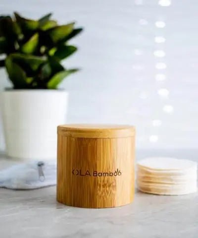 bamboo round box with "OLA Bamboo" engraved on the side. A pile of makeup remover pads, a mesh bag and a green plant in the background