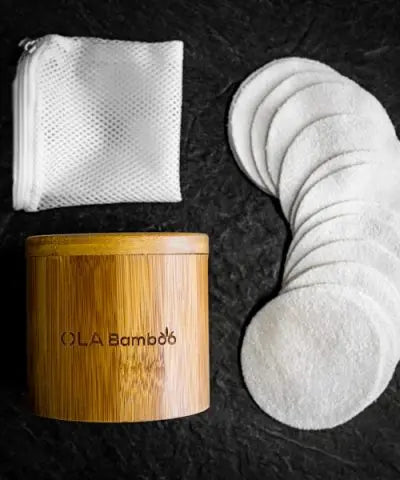 A mesh bag, a round bamboo box with "OLA Bamboo" engraved on the side, and a column of makeup remover pads set on a slate grey surface