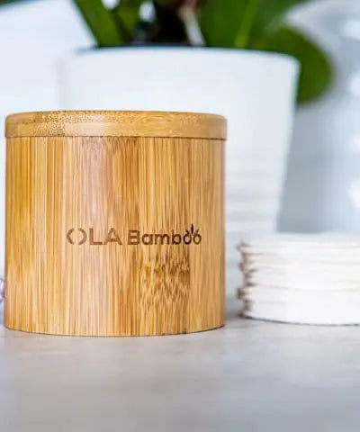 A round bamboo box with "OLA Bamboo" etched in the side, with a pile of makeup remover pads and a green plant in a white pot in the background