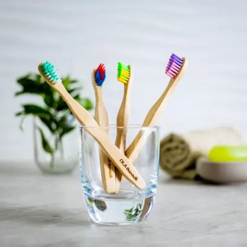 4 child's bamboo toothbrushes by Ola Bamboo set in a clear glass, with a towel, soap and a plant blurred in background