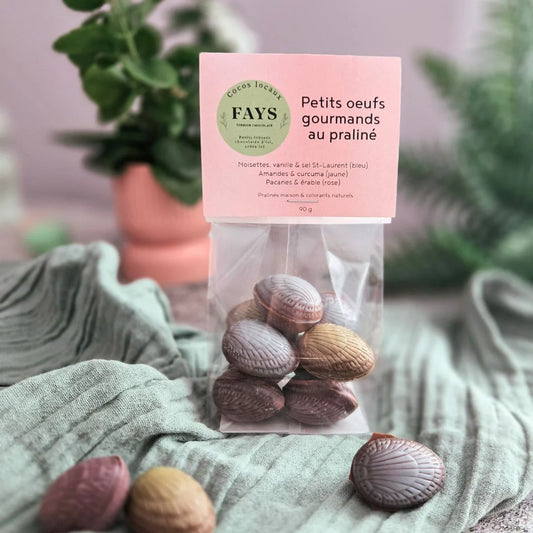 A clear bag of FAYS praline chocolate eggs labeled “Petits œufs gourmands au praliné” sits on a soft green cloth, with pastel-colored chocolate eggs inside and a blurred plant in the background.