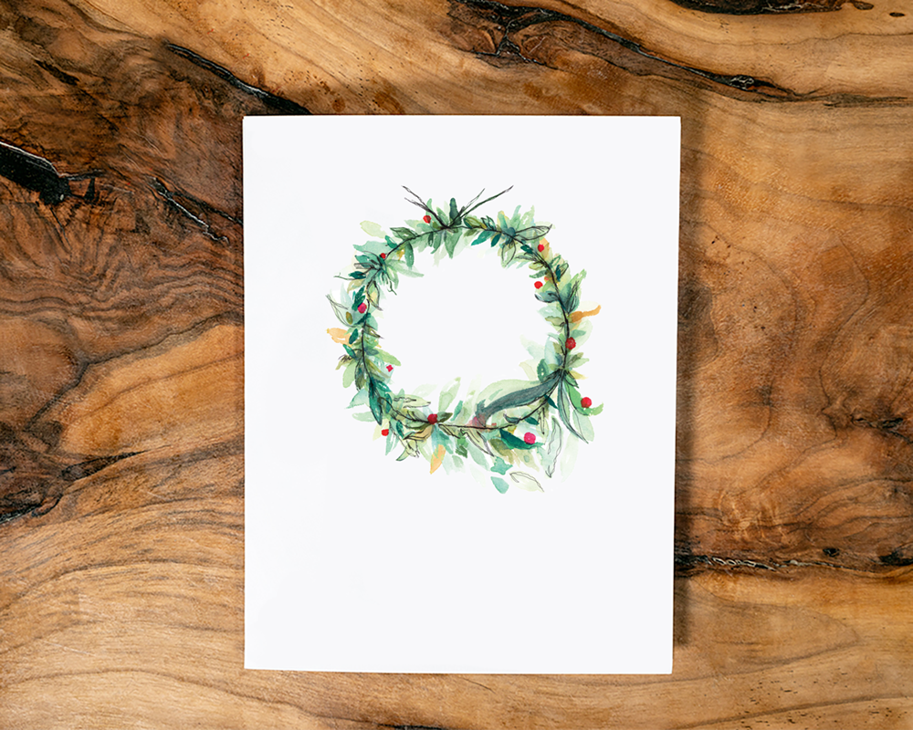 A watercolour design of a festive Christmas wreath on a white card placed on a wooden surface