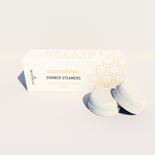 A box of "awaken" shower steamers sit on a pale background with two pale pucks (steamers) sit in front of box