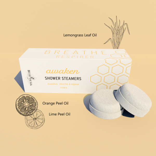 A box of "awaken" shower steamers on a pale yellow background with two steamers sitting in front. Ingredients are presented as sketches around the box