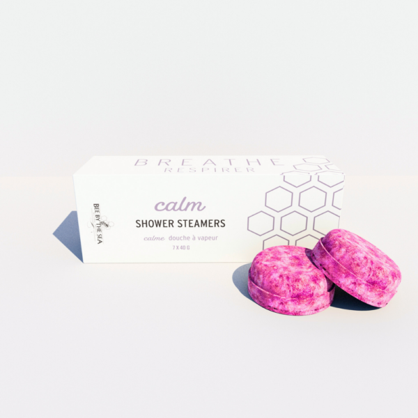 A box of "calm" shower steamers sit on a pale lavender background, with bright pink pucks sitting in front of box
