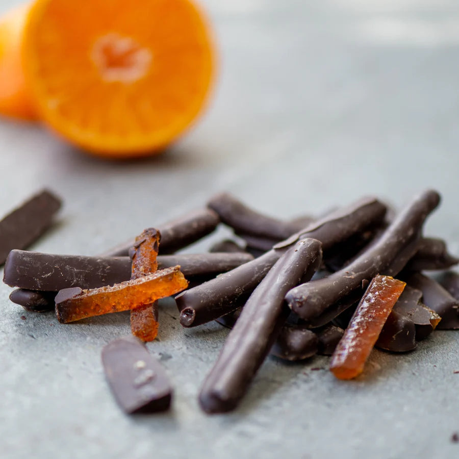 Chocolate-covered orange peels on a light surface with a halved orange in the background.