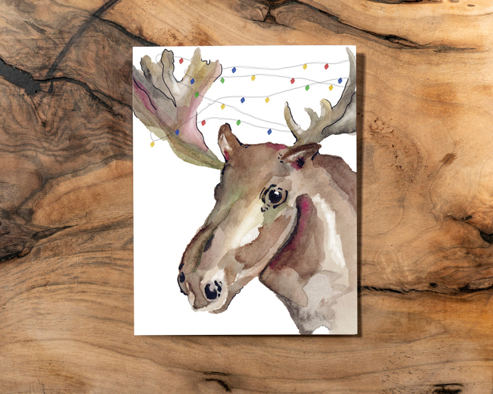 A watercolour illustration of a moose with antlers that are wrapped in Christmas lights on a white background, placed on a wooden surface.