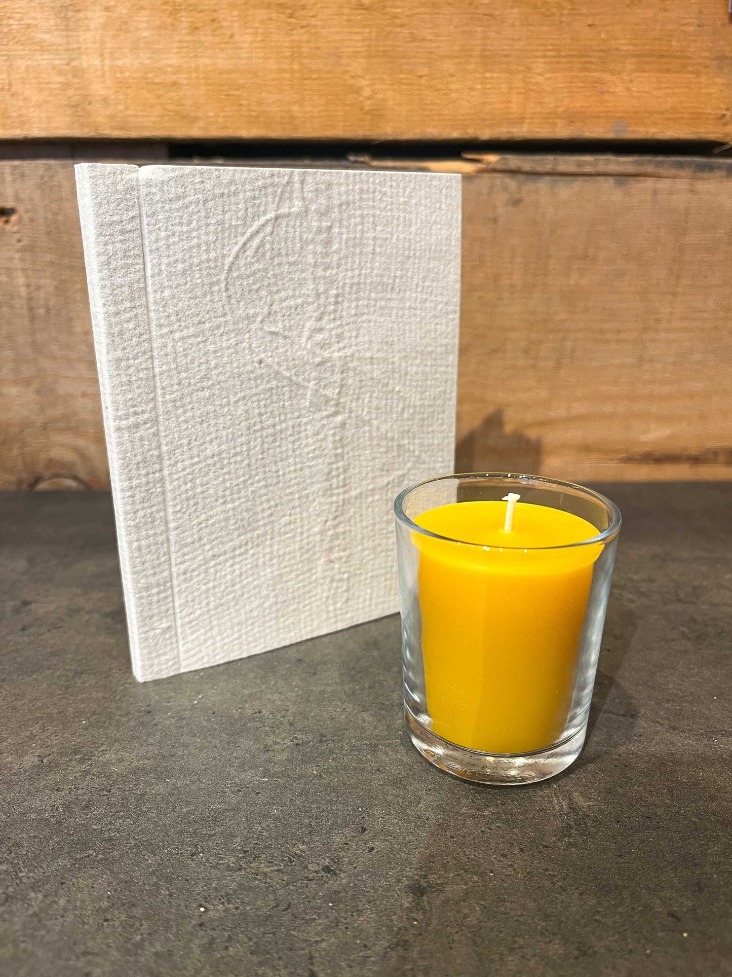 Handmade beeswax votive candle in glass cup next to beige eco-friendly notebook